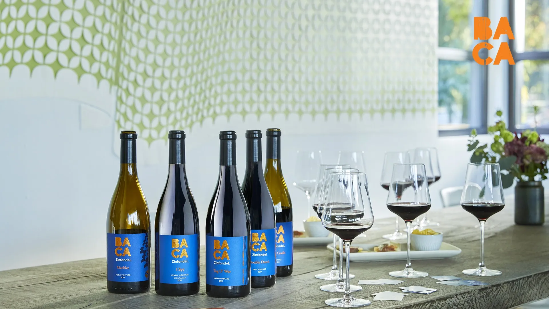 A row of BACA Zinfandel bottles and filled glasses on a white table