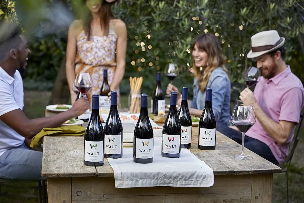 Friends laughing around an outdoor table set with a row of WALT bottles